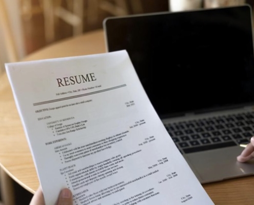 A person holding up a resume and looking at the screen of their laptop.