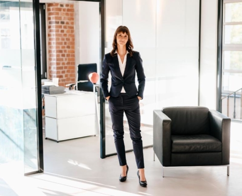 A woman in black suit and heels standing next to a chair.